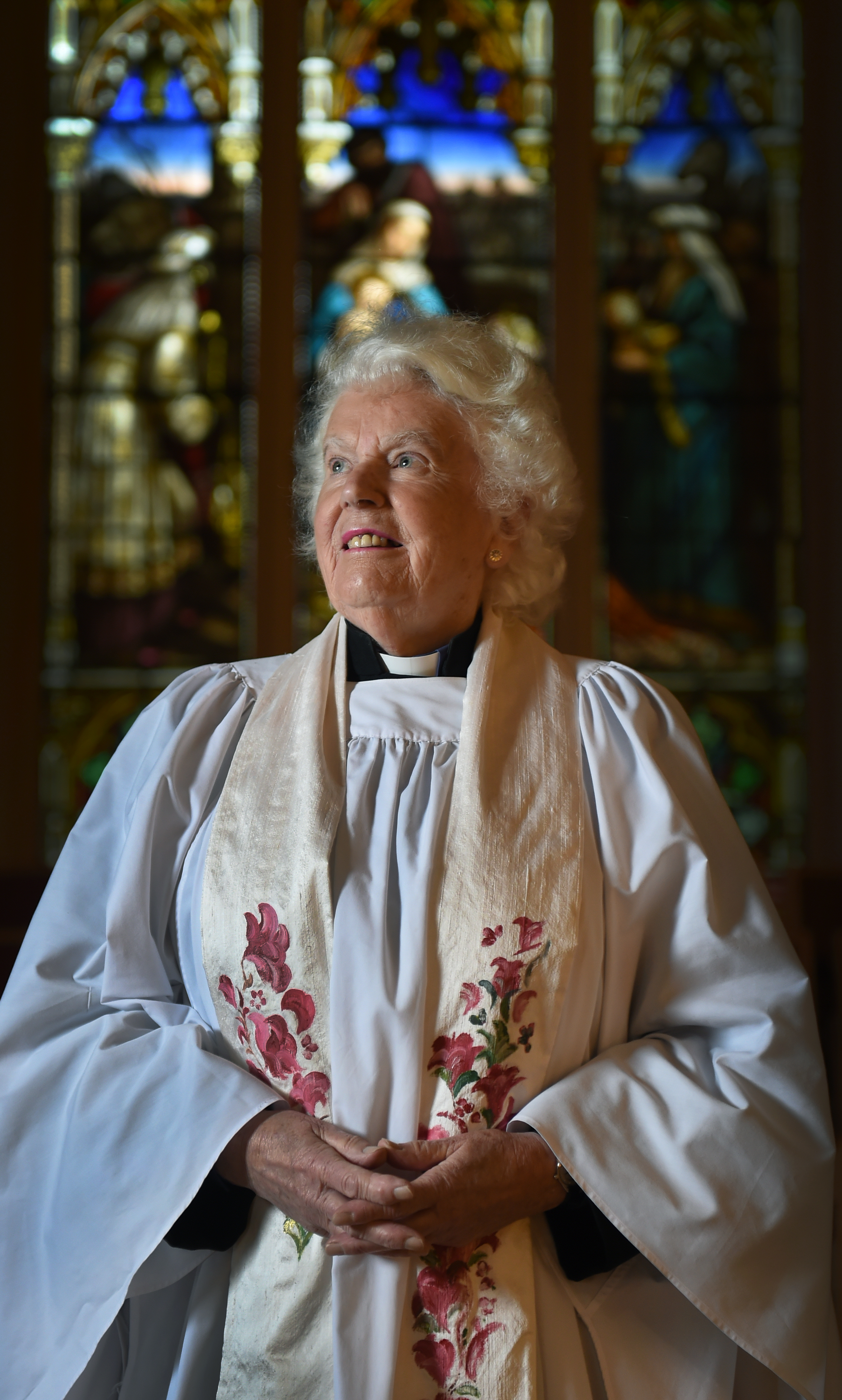 Awoman priest in her 90s with a stained glass window behind