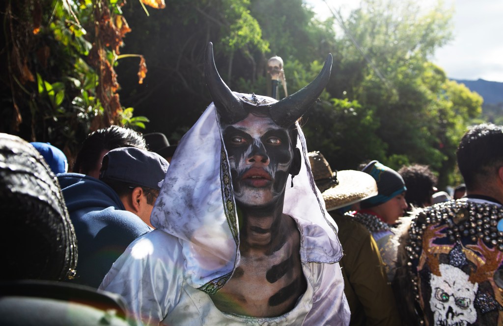 A man dressed up with black horns and a white hooded gown surronded by people looking in the opposite direction at an outdoor street festival