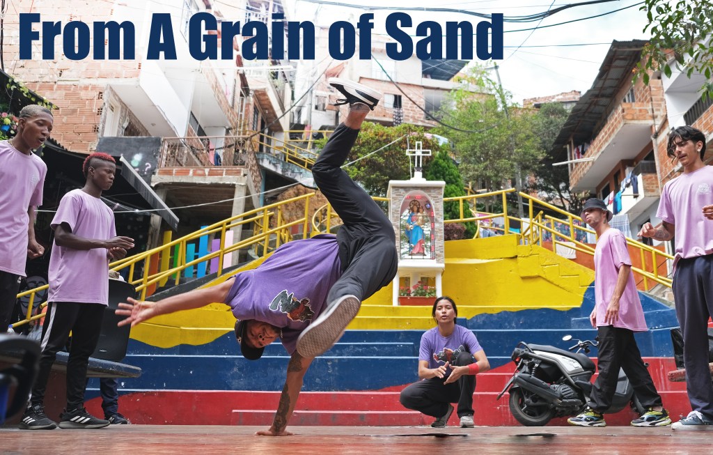 A poster image of a group of dancers in purple tshirts with one of them standing on one hand mid-dance move. Text says "From A Grain of Sand"